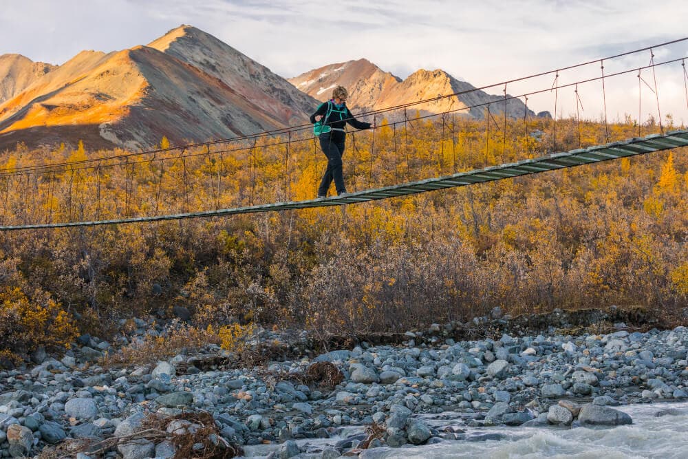 A person crosses a suspended bridge over a rocky river in a mountainous landscape during autumn.